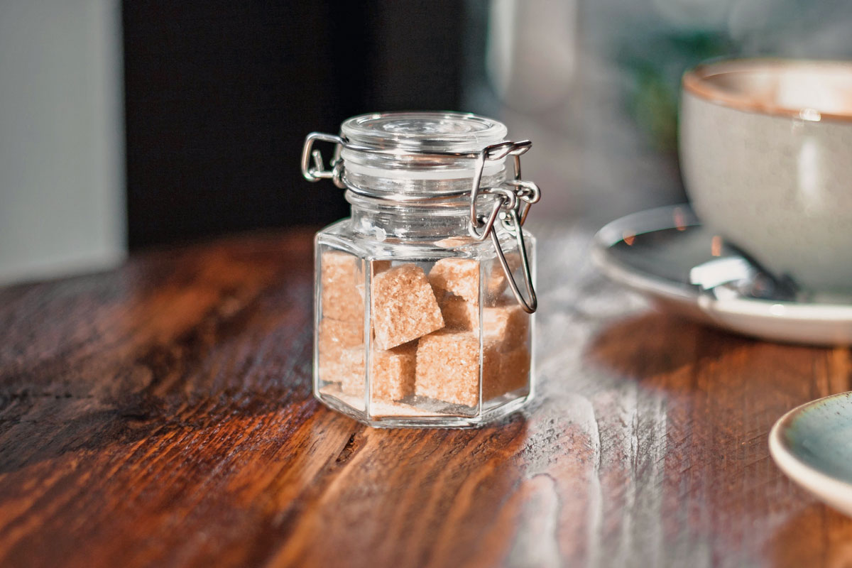 An image of a clear condiment shaker with brown sugar cubes near a gray teacup
