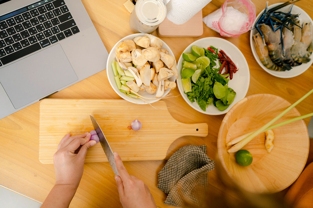 An aerial image of someone chopping food on a cutting board surrounded by small bowls of cut food and an open laptop