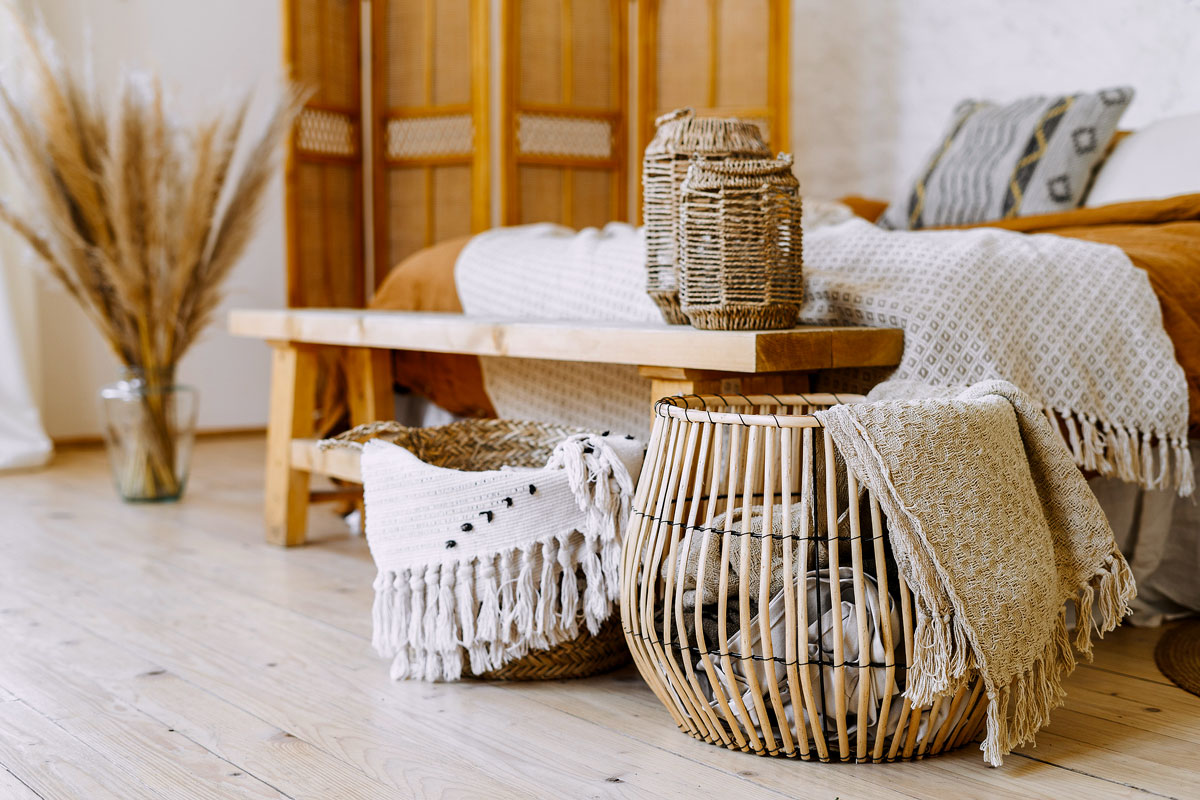 An image of a bohemian bedroom with textile sheet on bed, wooden bench seat, bamboo dressing screen, dry plants in vase, and wicker basket