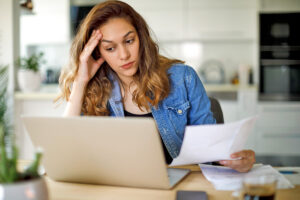 Woman with hand to forehead holding papers and looking at a computer